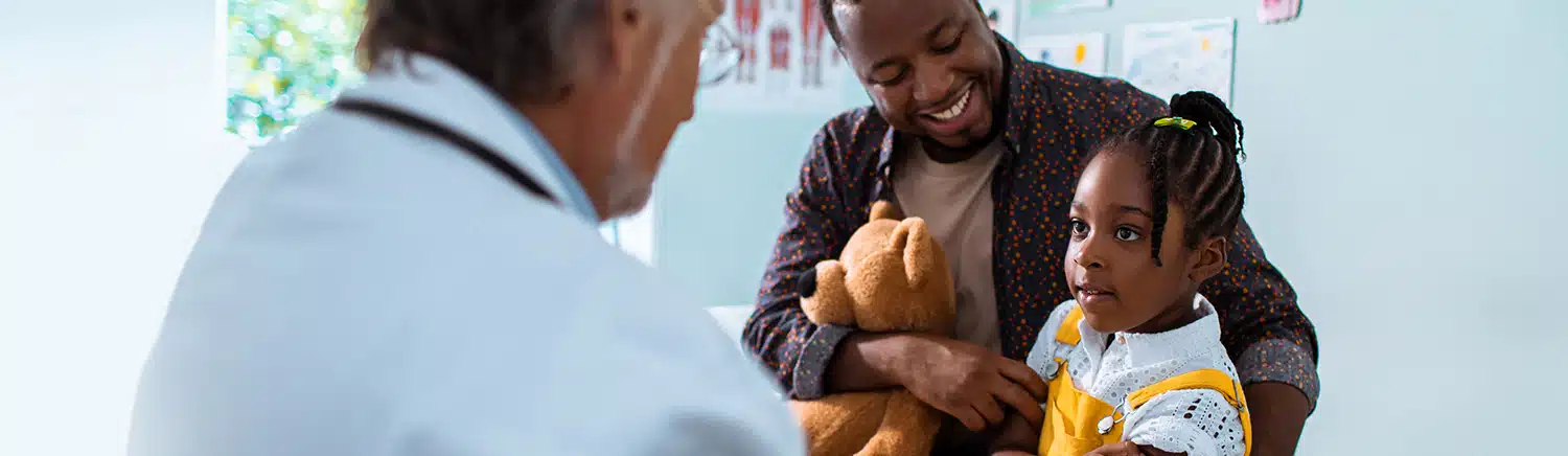 A father holding a stuffed animal in one arm and his daughter in the other consults with a doctor.